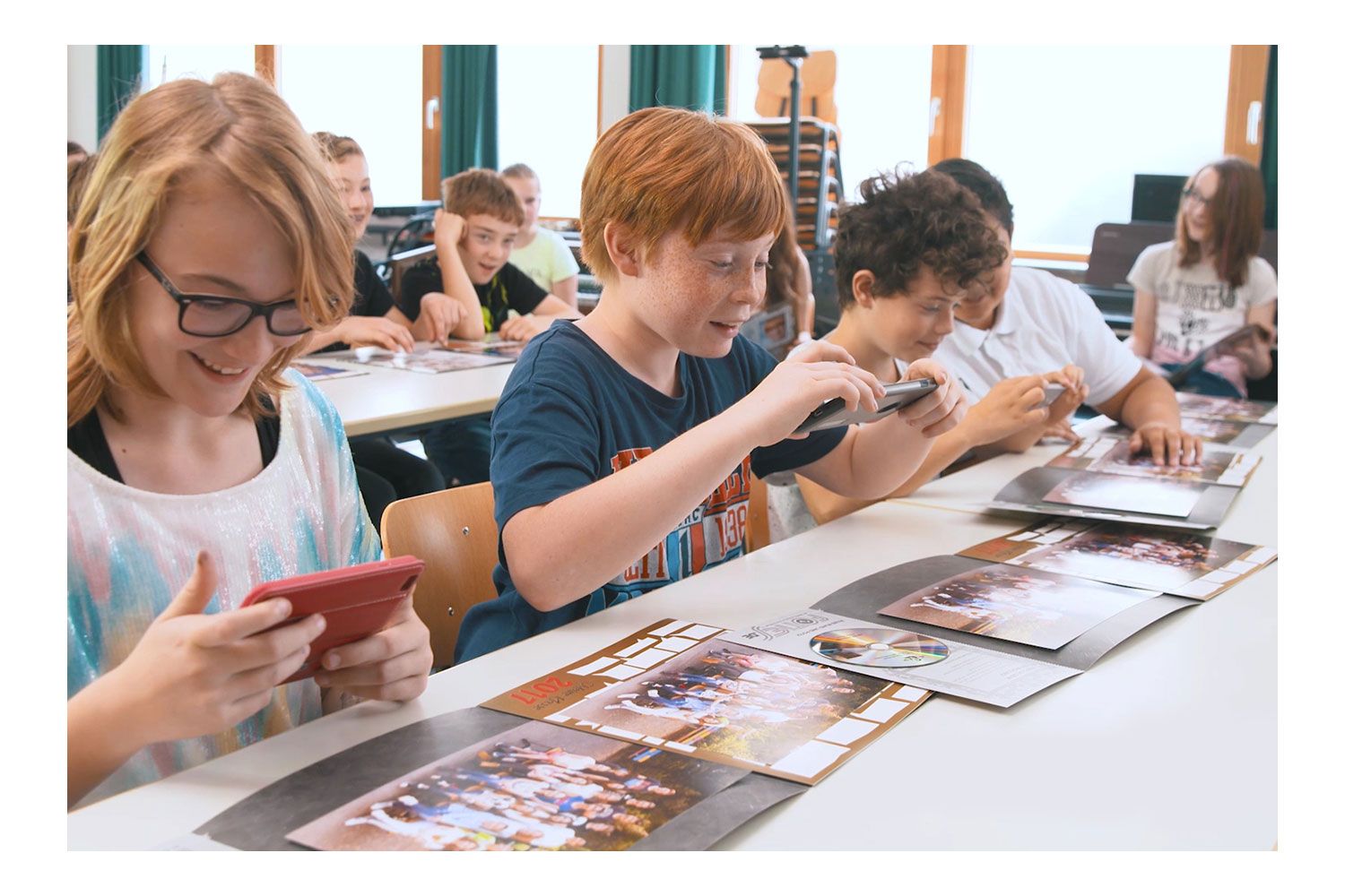 Schüler sitzen an ihren Plätzen und haben verschiedene Fotovariationen vor sich auf dem Tisch liegen, ein Junge macht ein Bild mit seinem Handy
